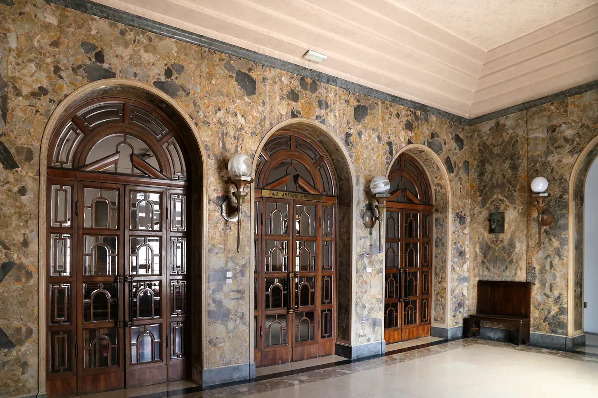 Three arched wooden doors with glass panels in the atrium, beside a wall of mottled stone and globe lamps
